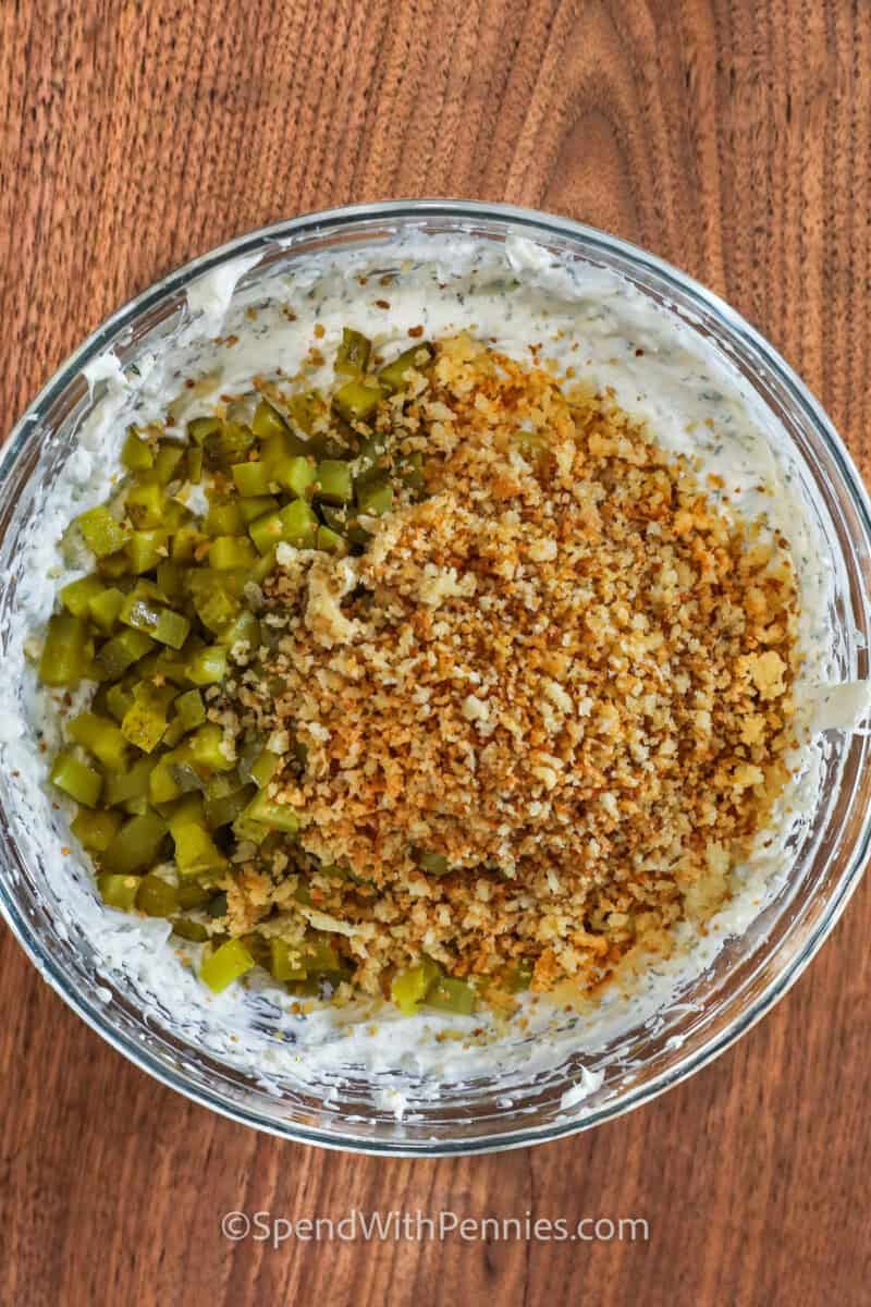 County fair-style deep-fried pickles are now a scrumptious fried pickle dip! Mix cream cheese, sour cream, diced pickles, savory spices, and crispy parmesan breadcrumbs for a crunchy, tangy treat. Add flavors like ranch or buffalo to customize it. With pantry ingredients, this dip offers an irresistible taste of the south. #spendwithpennies adding pickles and breadcrumbs to bowl to make Fried Pickle Dip