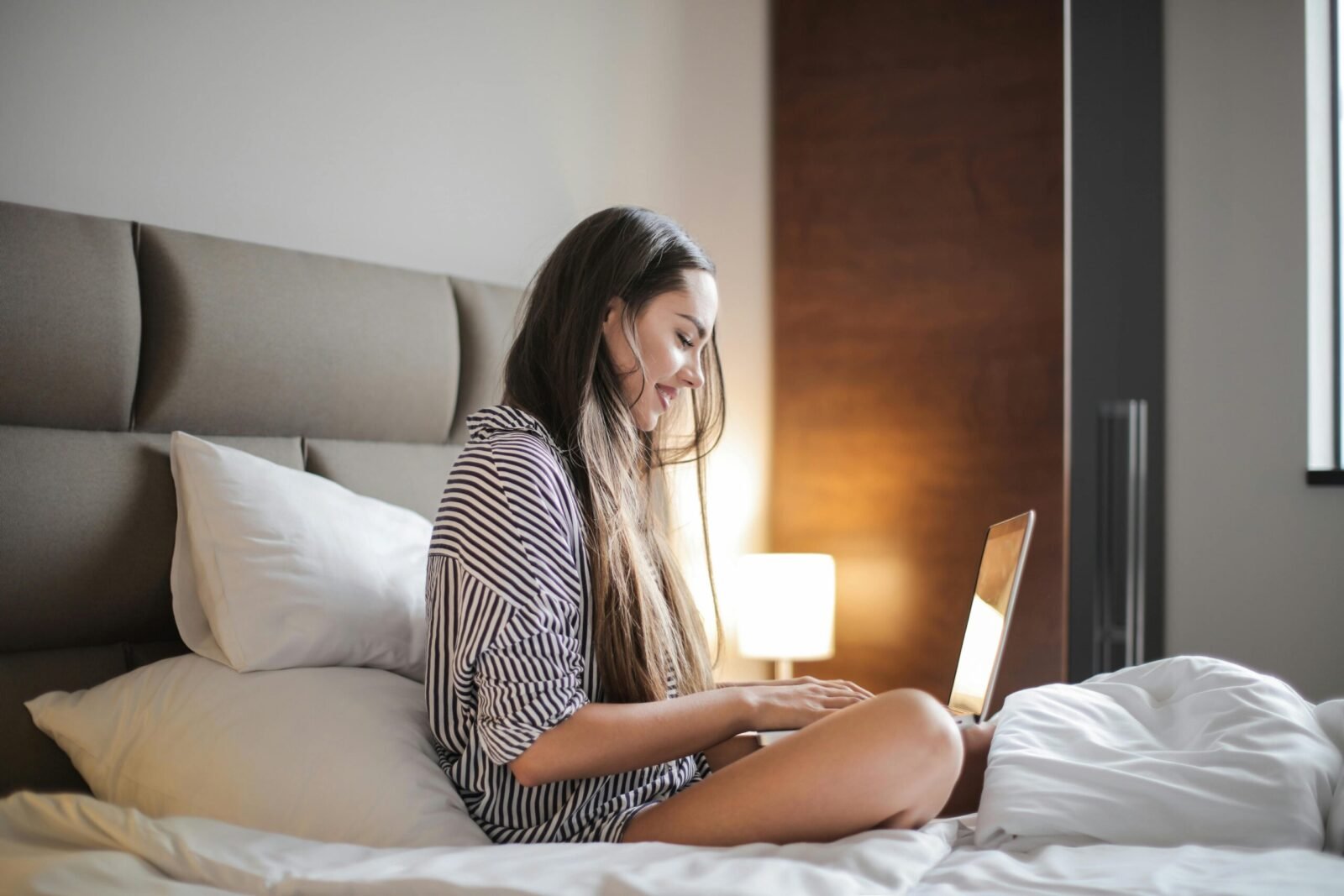 side view photo of smiling woman in a black and white striped top sitting on a bed while using a lap