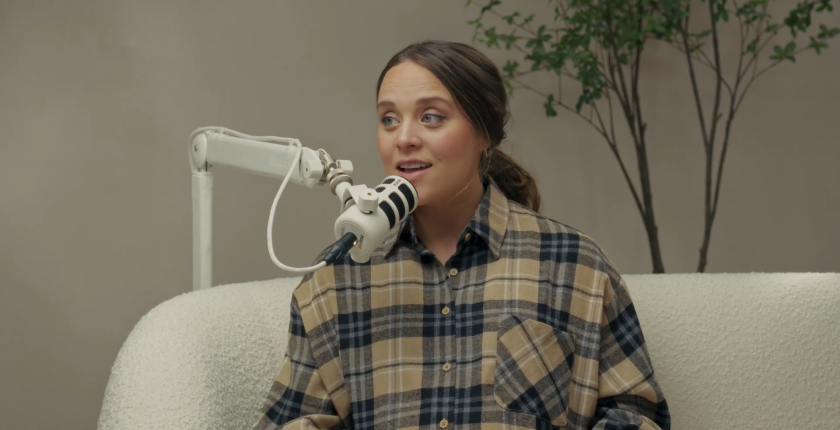 Jinger Duggar speaks into a mic while wearing a beige plaid shirt.