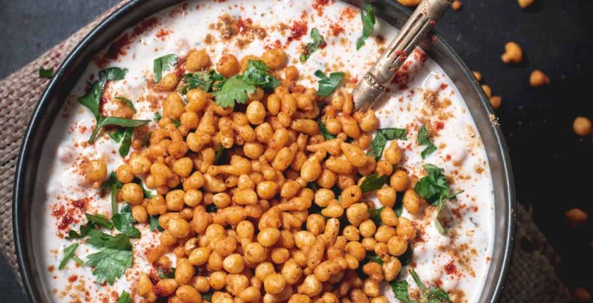 An overshot of boondi raita garnished with cilantro and served in a black bowl with a golden spoon.