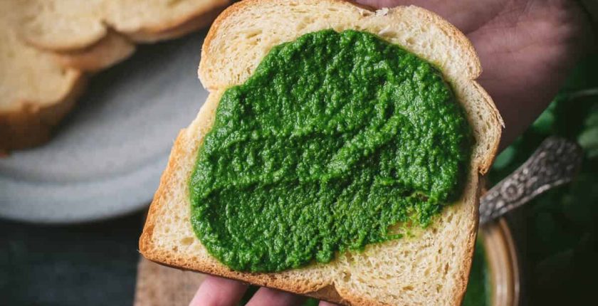 Bread slathered with green chutney is placed on a palm. There are additional bread slices kept on a grey plate.